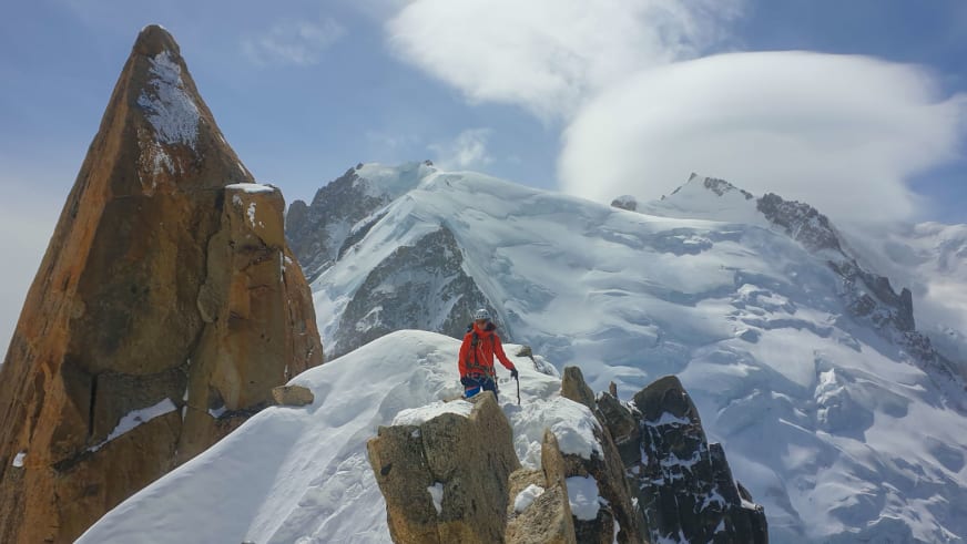 Bergwanderführer Christoph Knappe in roter Winterjacke steht auf schneebedecktem Felsgrat mit Bergstock zwischen Felsturm und Gletscher.