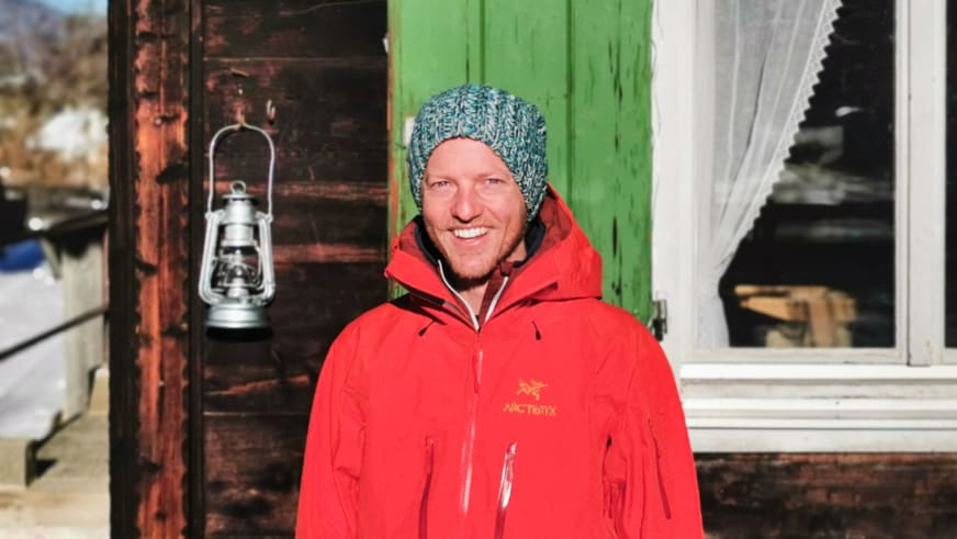 Bergwanderführer Christoph Knappe mit grau-blauer Strickmütze und roter Arc'teryx Jacke lächelt vor einer Holzhütte mit Petroleumlaterne.