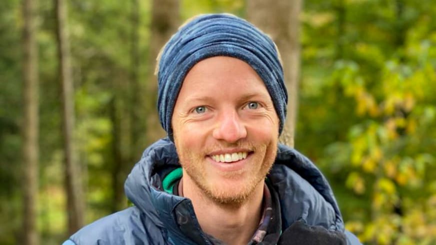 Portrait von Bergwanderführer Christoph Knappe mit blauer Strickmütze und dunkler Softshelljacke vor unscharfem Laubwald bei Tageslicht.