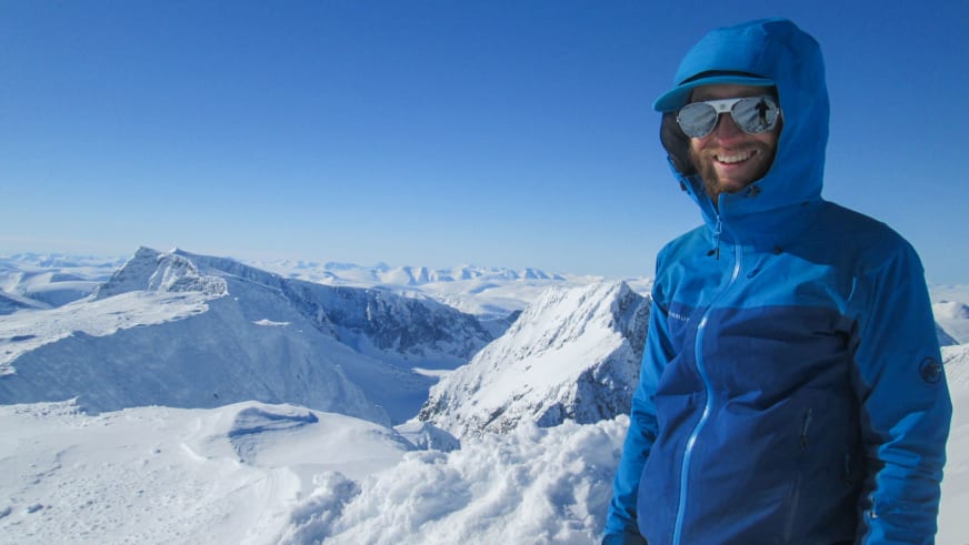 Bergwanderführer Bas Pijpers lächelt in blauer Hardshelljacke auf schneebedecktem Berggipfel mit Hochgebirgspanorama.