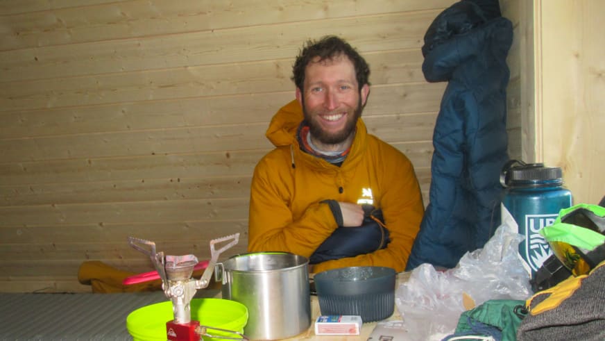 In einer Berghütte sitzt Bergwanderführer Bas Pijpers in gelber Daunenjacke mit Kochausrüstung auf dem Boden.