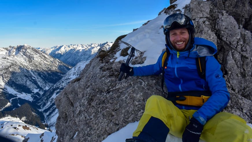 Bergwanderführer Bas Pijpers sitzt lächelnd in blauer Winterjacke mit Eispickel auf verschneitem Felsgrat.