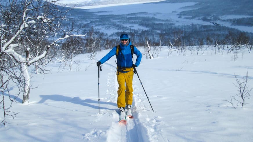 Bergwanderführer Bas Pijpers steht lächelnd auf Skiern im tiefen Pulverschnee zwischen schneebedeckten Birken.