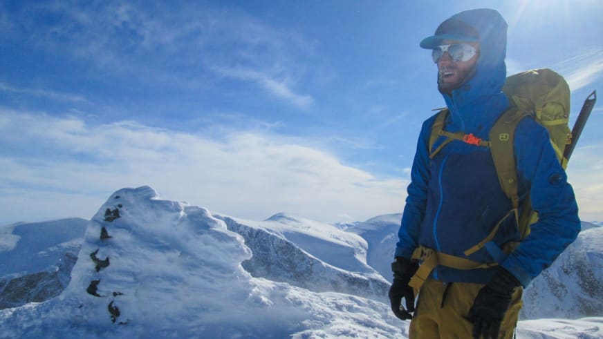 Auf schneebedecktem Berggipfel steht Bergwanderführer Bas Pijpers in blauer Winterjacke mit grünem Rucksack.