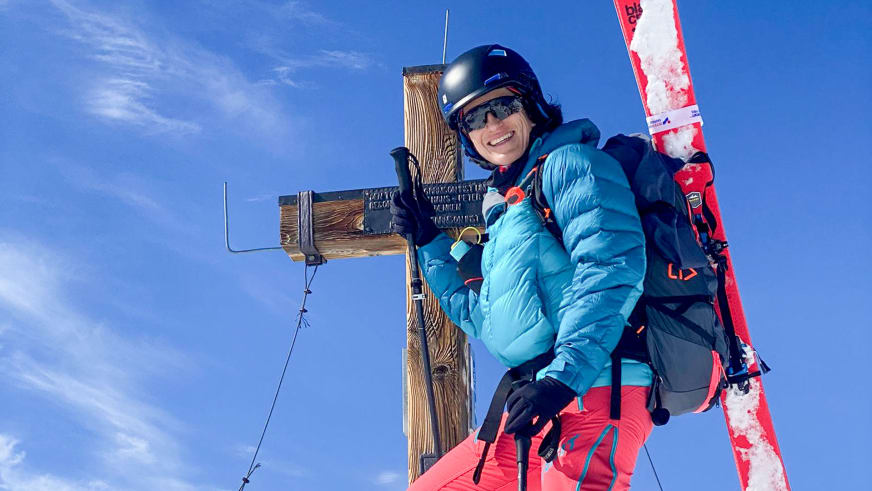 Am hölzernen Gipfelkreuz lächelt Bergwanderführerin Alexandra Querl in türkiser Daunenjacke und roter Skihose.