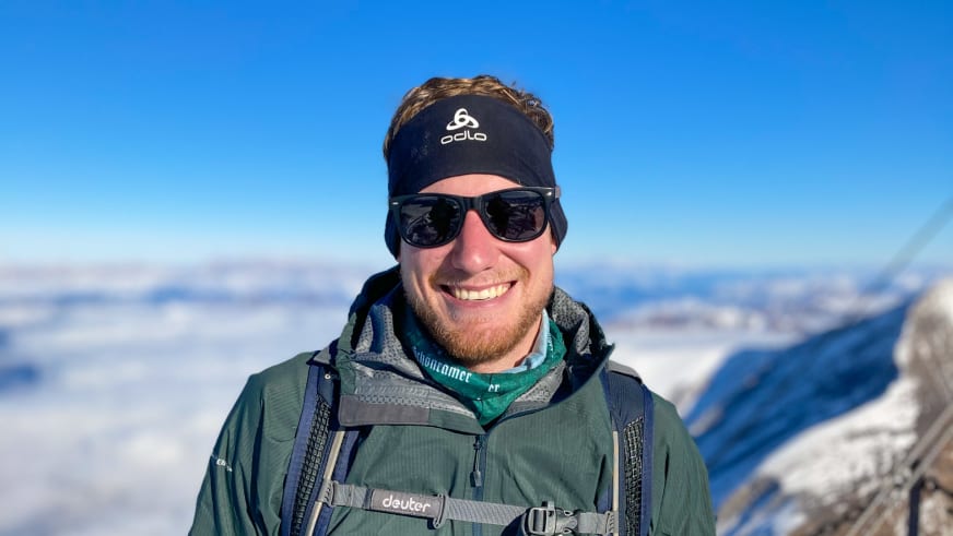 Bergführer Simon Deckenbrock lächelt mit Sonnenbrille und grüner Jacke vor schneebedeckten Berggipfeln.