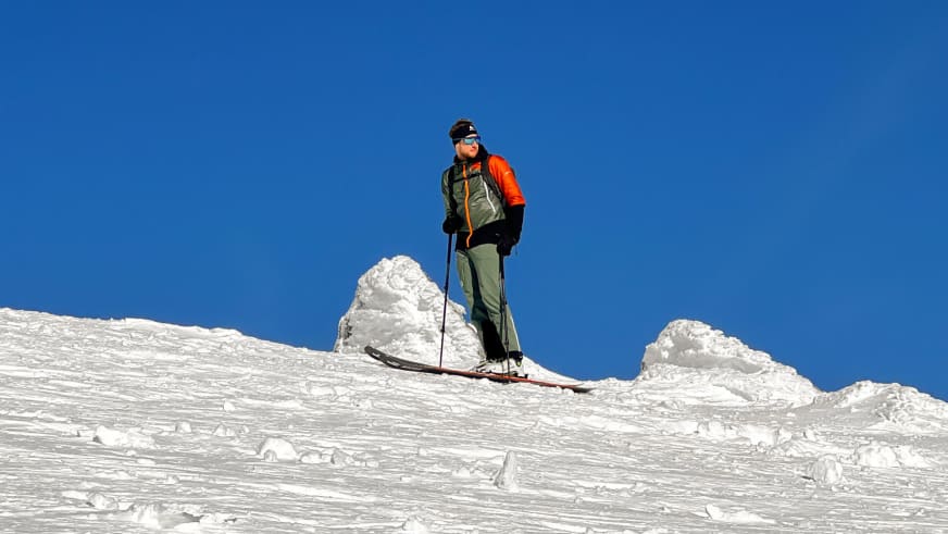 Bergführer Simon Deckenbrock steht auf Tourenski in orange-schwarzer Jacke vor intensiv blauem Himmel.
