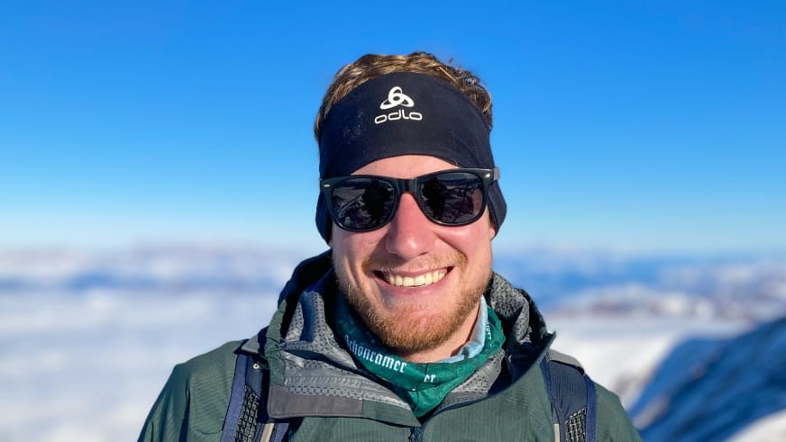 Bergführer Simon Deckenbrock mit schwarzer Stirnband-Mütze und olivgrüner Jacke steht vor schneebedeckten Gipfeln.