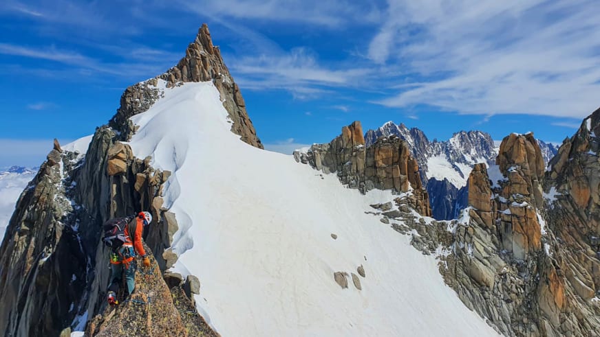 Bergführer Simon Deckenbrock klettert in oranger Jacke an steiler Felswand zwischen schneebedeckten Gipfeln.