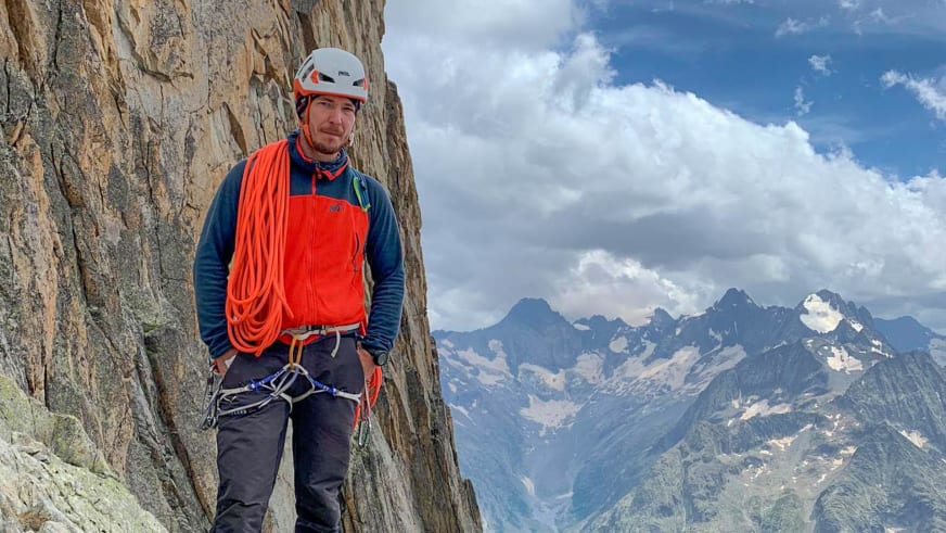 An einer senkrechten Felswand steht Bergführer Márk Salamon mit orange Kletterweste und aufgerolltem Seil.