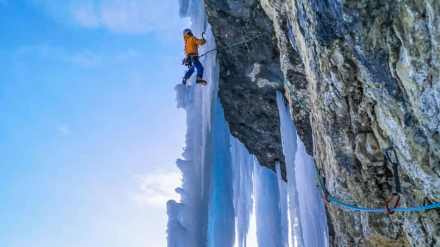 Bergführer Tobias Riedlsperger klettert in oranger Winterjacke an einer steilen Eiswand mit blauen Eiszapfen.