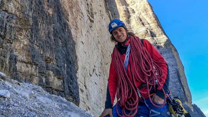 Bergführer Tobias Riedlsperger trägt rote Kletterjacke und blauen Helm, mit rotem Kletterseil um die Schultern vor Kalkstein-Felswand.