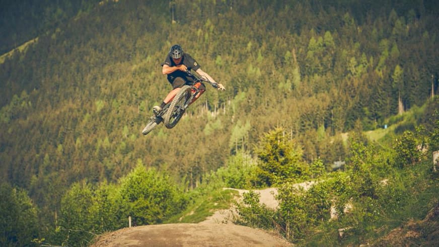 Bergführer Tobias Riedlsperger führt einen Mountainbike-Sprung über eine Erdaufschüttung im bewaldeten Gelände aus.