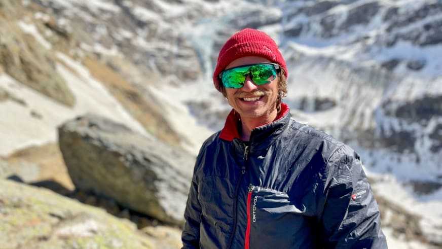 Mit roter Strickmütze und grüner Sonnenbrille lächelt Bergführer Tobias Riedlsperger vor steiniger Berglandschaft.