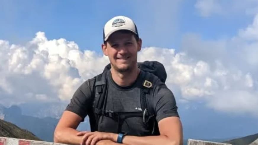 Bergwanderführer Julian Götz sitzt entspannt vor einer Berglandschaft mit weißer Kappe und grauem T-Shirt.