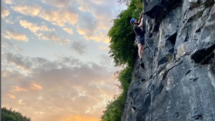 An einer steilen Felswand klettert Bergwanderführer Julian Götz mit blauem Helm und weißem Kletterseil bei Abendstimmung.