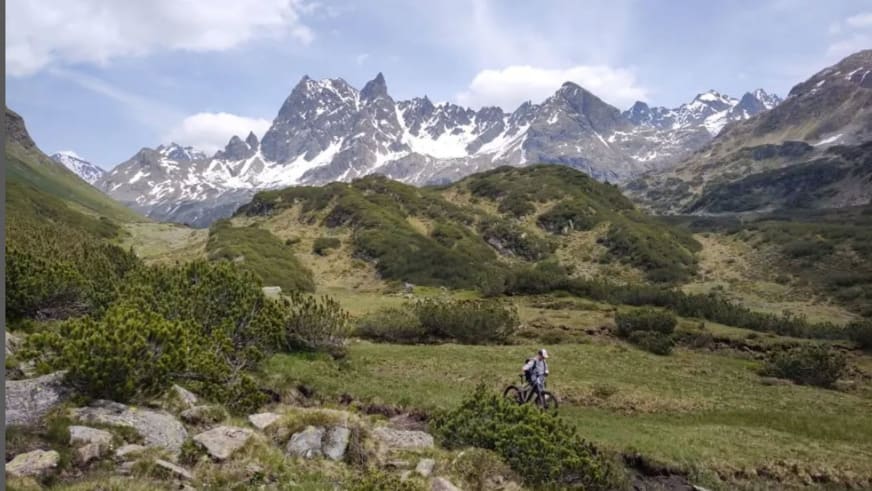 Bergwanderführer Julian Götz steht mit seinem Mountainbike auf grüner Wiese vor schneebedeckten Bergen im alpinen Tal.