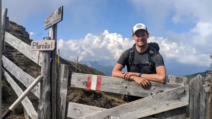 Bergwanderführer Julian Götz lehnt lächelnd an einem Weidezaun neben einem Holzwegweiser zum Pfannlörl.