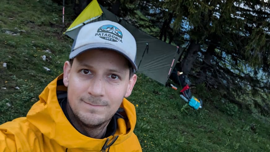 Bergwanderführer Julian Götz macht ein Selfie vor seinem grauen Zelt zwischen Nadelbäumen auf einer Wiese.