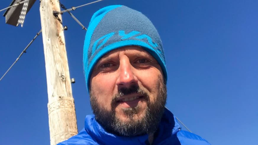 Am Gipfelkreuz lächelt Bergwanderführer Thomas Ziegler in blauer Wintermütze und Daunenjacke in die Kamera.