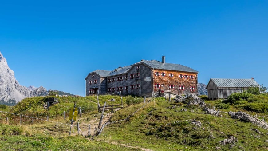 Außenansicht der Hofpürglhütte mit traditionellen Holzbauten vor der Bergkulisse des Dachsteinmassivs.
