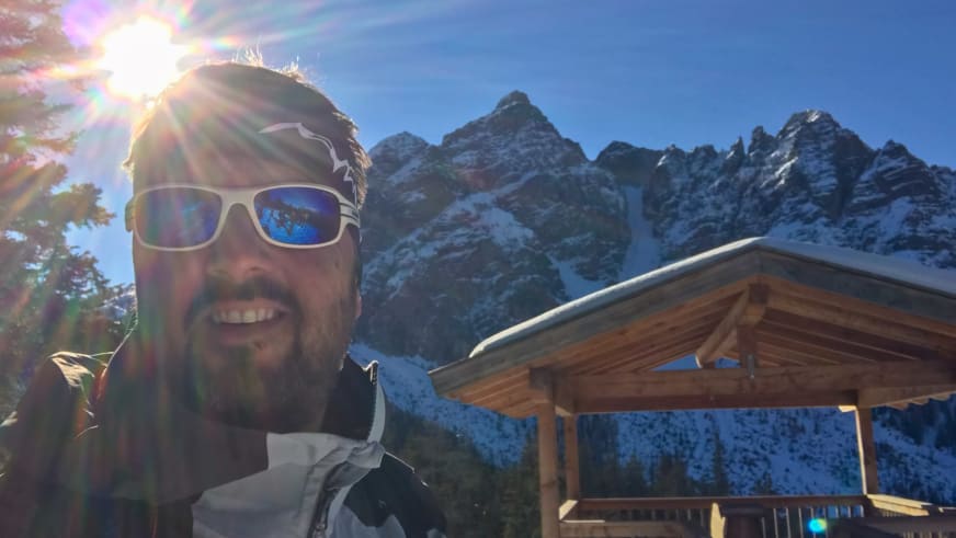 Bergwanderführer Thomas Ziegler mit Sonnenbrille vor schneebedeckter Bergkette und traditionellem Holzgebäude im Winter.