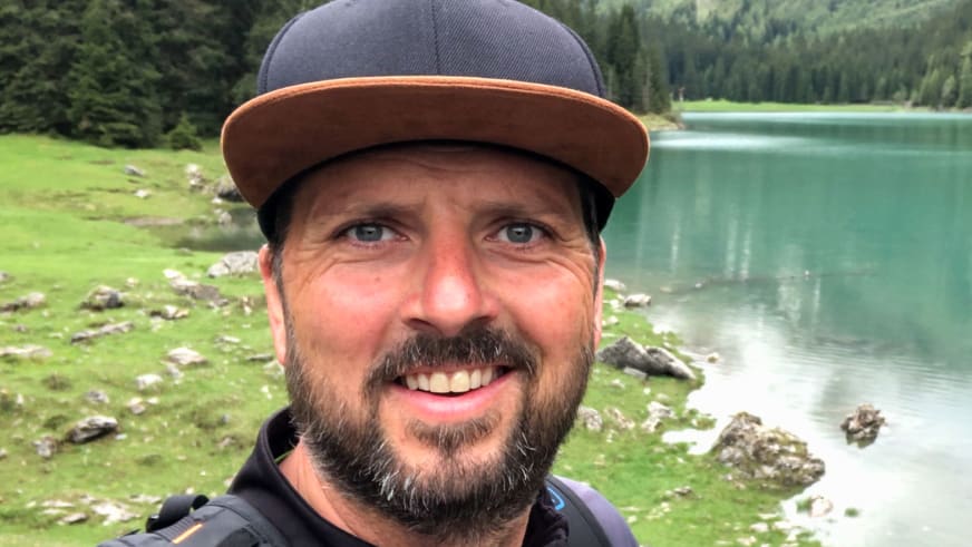 Portrait von Bergwanderführer Thomas Ziegler mit grauer Kappe vor einem türkisen Bergsee mit grünen Almwiesen und Nadelwald.