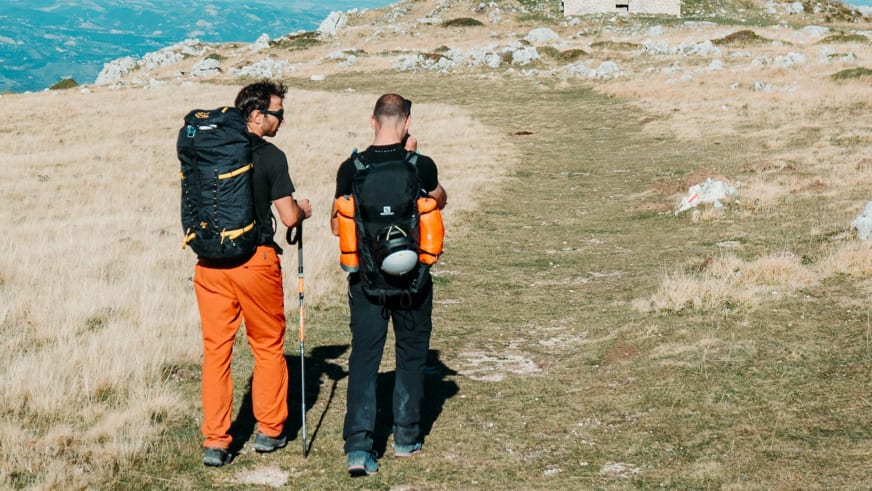 Zwei Personen mit Rucksäcken stehen auf grasigem Plateau und blicken über alpine Hochebene - Bergführer Enrico Trapazzo führt.