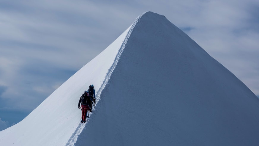 Zwei angeseilte Bergsteiger steigen auf einem schmalen Schneegrat zu einem pyramidenförmigen Gipfel auf - Bergführer Enrico Trapazzo führt die Tour.