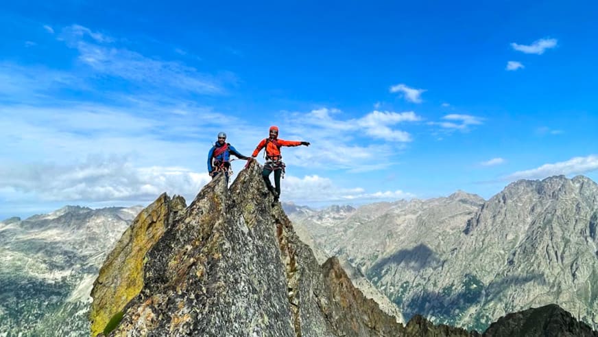 Zwei Personen mit ausgebreiteten Armen auf spitzem Berggipfel mit Helmen und Klettergurten vor Bergpanorama - Bergführer Enrico Trapazzo.