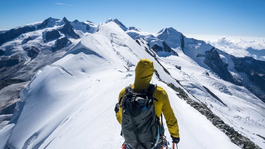 Person in gelber Daunenjacke steht auf schmalem Schneegrat und blickt auf vergletscherte Berge - Bergführer Enrico Trapazzo führt.