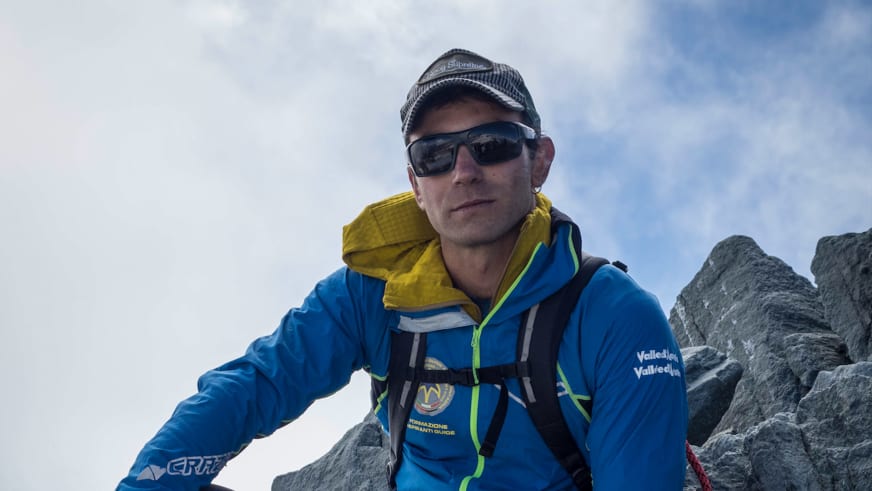 Bergführer Enrico Trapazzo sitzt entspannt in blauer Softshelljacke und Sonnenbrille vor grauem Felsgestein.