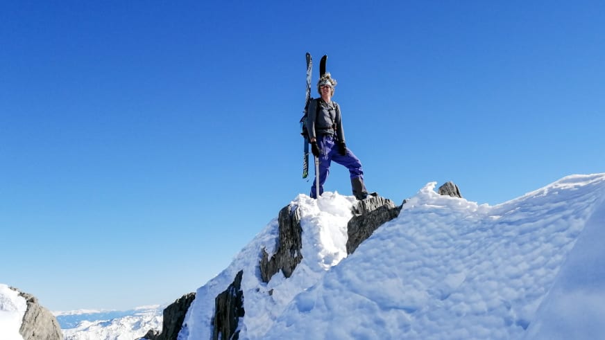 Bergführer Fabian Bendlin hält zwei Tourenskier in die Höhe auf schneebedecktem Felsgipfel.