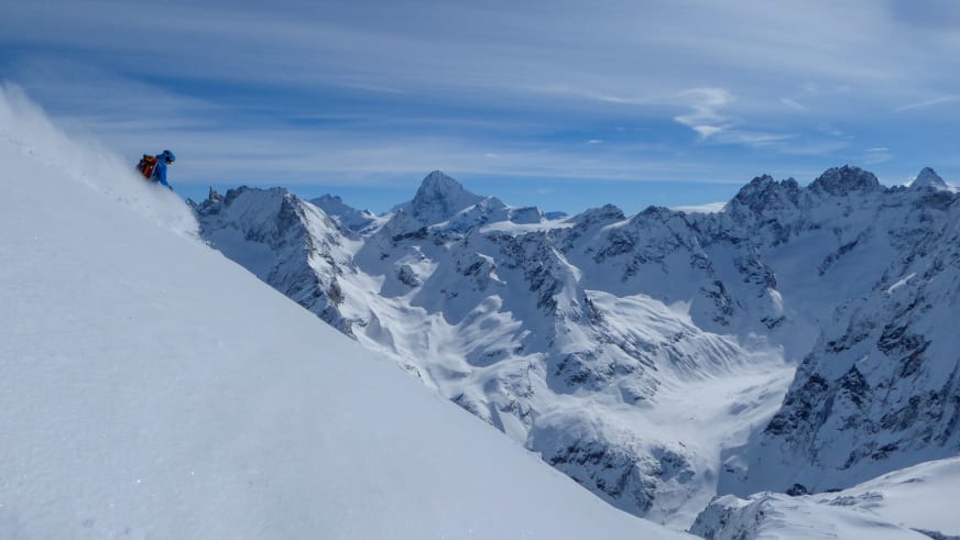 Bergführer Fabian Bendlin fährt in blauer Jacke einen steilen Schneehang mit Hochgebirgspanorama hinab.