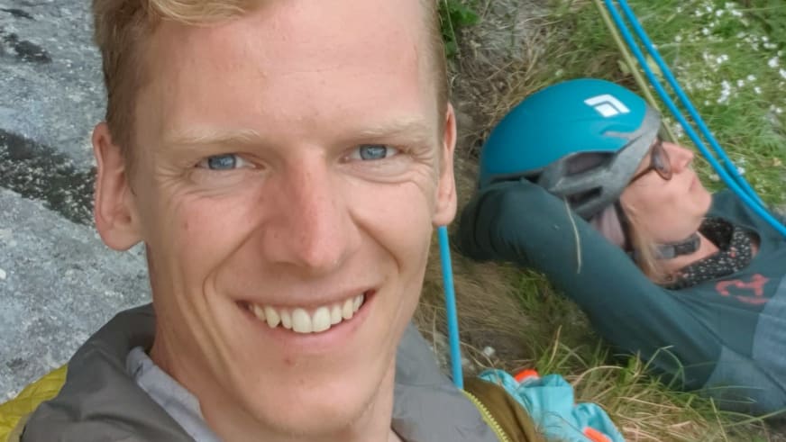 Lächelndes Portrait von Bergführer Fabian Bendlin in grauer Jacke mit zweiter Person im Hintergrund am Fels.