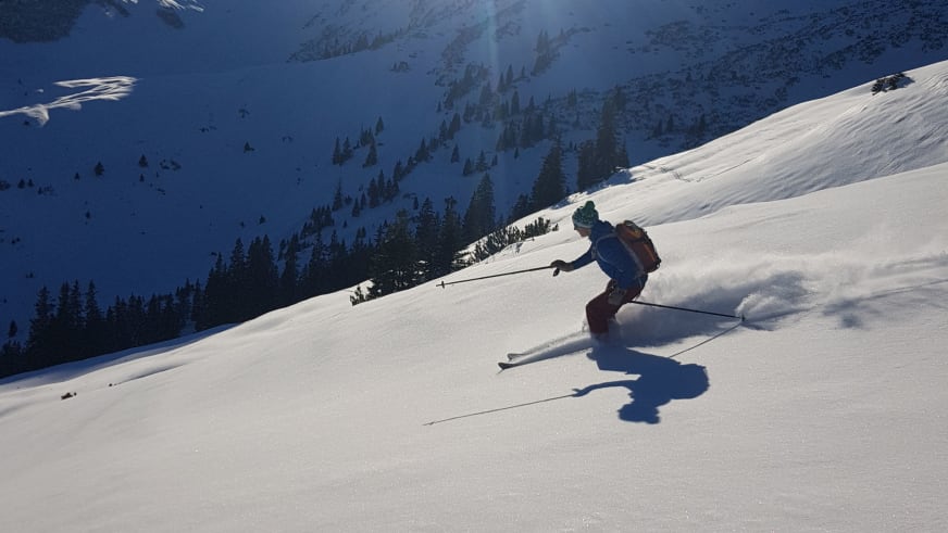 Bergführer Fabian Bendlin in grünem Helm fährt einen Schneehang mit Pulverschnee-Aufwirbelung hinab.