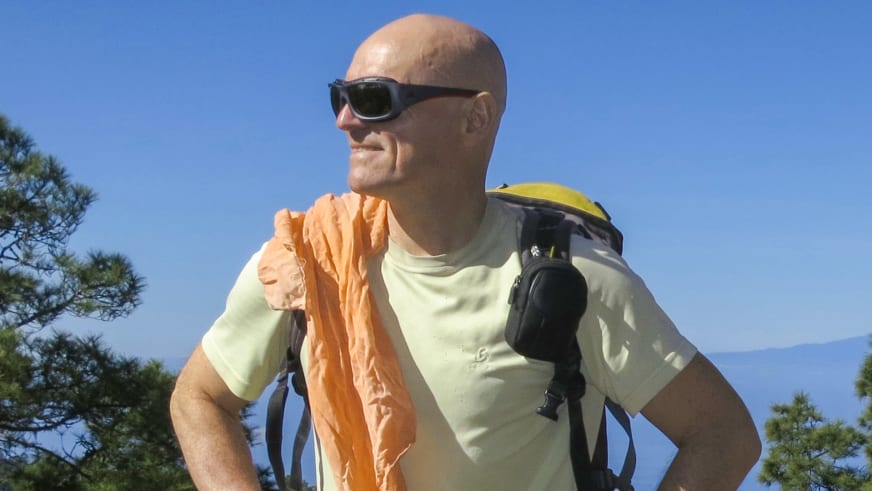 Bergwanderführer Reinald Zölch mit schwarzer Sonnenbrille und grauem Rucksack vor grünen Baumwipfeln.