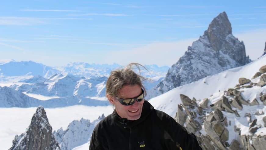 Bergführer Franz Hölzl mit Sonnenbrille und schwarzer Softshell-Jacke vor pyramidenförmigem Felsgipfel im Hochgebirge.