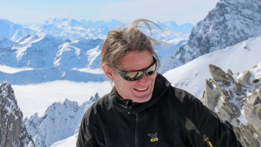 Bergführer Franz Hölzl mit Sonnenbrille und schwarzer Bergjacke lächelt vor hochalpiner Winterlandschaft mit schneebedeckten Bergen.