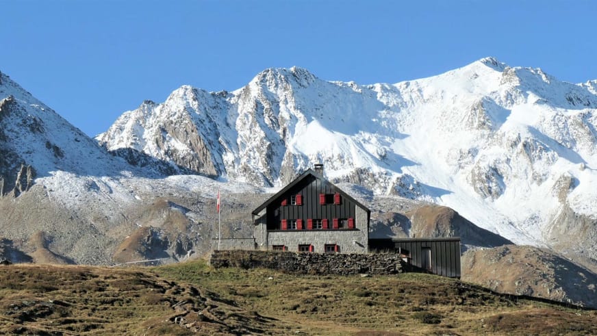 Die Vermigelhütte mit gehisster Schweizer Flagge vor schneebedeckten Bergen der Gotthardgruppe im Herbst.