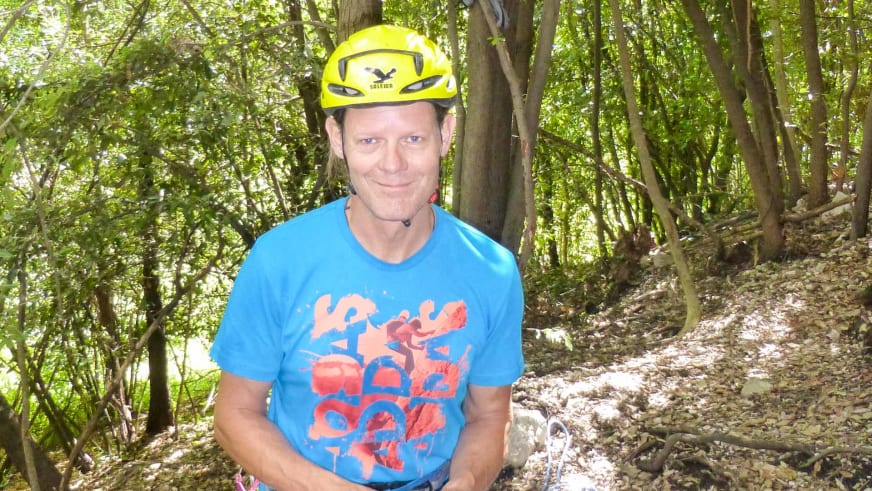 Bergführer Franz Hölzl trägt einen gelben Salewa-Kletterhelm und blaues T-Shirt im Waldgebiet mit dichtem Laubwald im Hintergrund.
