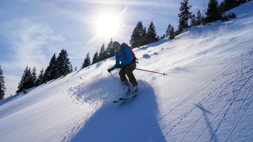 In blauer Jacke und grünem Helm fährt Bergführer Franz Hölzl eine steile Schneeflanke hinab und wirbelt Pulverschnee auf.