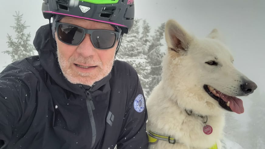Bergführer Franz Hölzl mit weißem Bart und Sonnenbrille neben einem weißen Hund mit gelbem Halsband im verschneiten Wald.