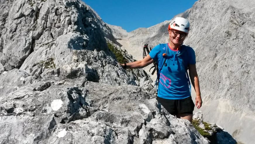 Zwischen hellen Kalksteinfelsen steht Bergwanderführerin Simone Thater mit weißem Helm und Wanderstöcken vor Bergkulisse.