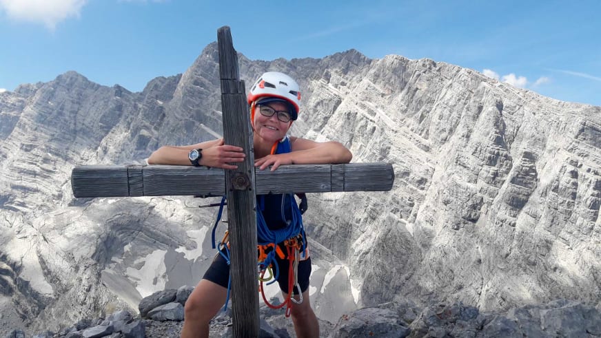 Bergwanderführerin Simone Thater steht mit weißem Helm und Klettergurt am hölzernen Gipfelkreuz vor Kalkstein-Berglandschaft.