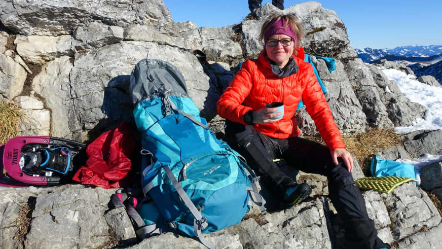 Bergwanderführerin Simone Thater sitzt mit Tasse auf Kalkstein und macht Pause neben roten Schneeschuhen und blauem Rucksack.