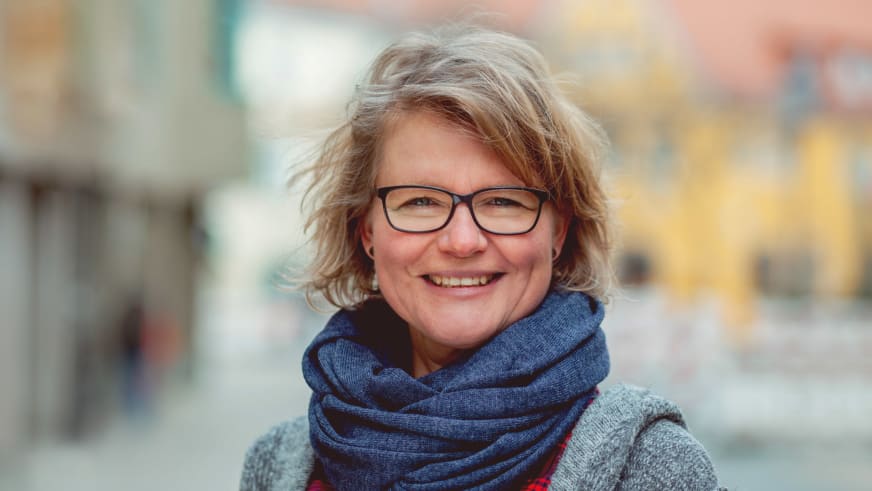 Bergwanderführerin Simone Thater mit blonden Haaren und schwarzer Brille trägt blauen Schal vor städtischer Kulisse.