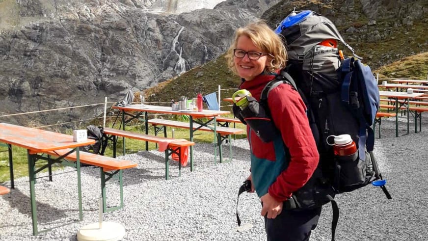 Bergwanderführerin Simone Thater mit blauem Rucksack und roter Fleecejacke vor Almhütte mit Wasserfall im Hintergrund.