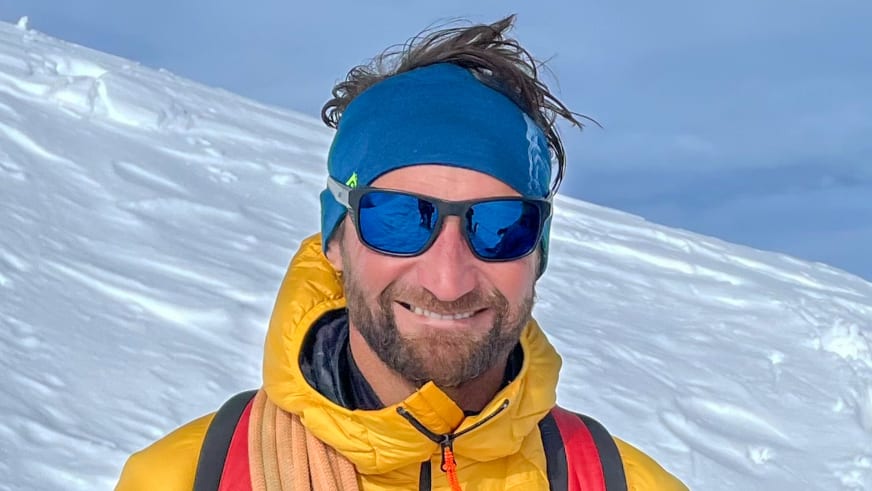 Bergführer Klaus Petz in gelber Daunenjacke mit verspiegelter Sonnenbrille vor verschneiter Berglandschaft bei klarem Sonnenlicht.