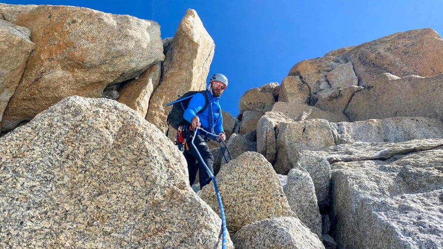 Bergführer Klaus Petz steht mit blauem Kletterseil zwischen großen Granitfelsblöcken und trägt blaue Kletterjacke sowie weißen Helm.